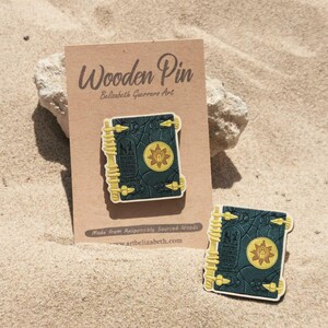 K&ouml;nnte beinhalten: Holz-Anstecknadel mit detailliertem Buchdesign mit dunkelgr&uuml;nem Einband, goldenen Akzenten und einem zentralen Emblem. Die Anstecknadel wird auf einer braunen Karte mit dem Text "Wooden Pin" und "Belisabeth Guerreros Art" pr&auml;sentiert. Eine weitere Anstecknadel ist ebenfalls sichtbar.