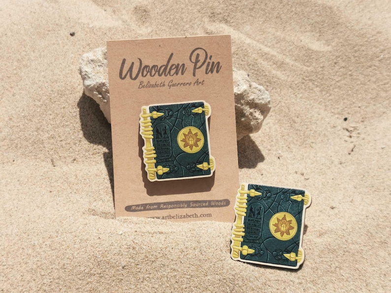 K&ouml;nnte beinhalten: Holz-Anstecknadel mit detailliertem Buchdesign mit dunkelgr&uuml;nem Einband, goldenen Akzenten und einem zentralen Emblem. Die Anstecknadel wird auf einer braunen Karte mit dem Text "Wooden Pin" und "Belisabeth Guerreros Art" pr&auml;sentiert. Eine weitere Anstecknadel ist ebenfalls sichtbar.