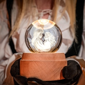May include: A glowing crystal ball with a carved image of two figures, resting on a wooden base. The sphere is held by a person wearing a white top. The figures inside the sphere appear to be embracing. The wooden base is cylindrical.