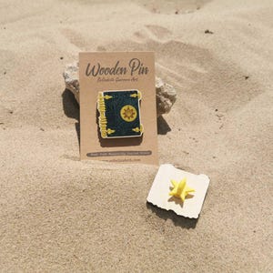 K&ouml;nnte beinhalten: Ein Holz-Pin in Form eines Buches mit goldenem Sonnendesign, pr&auml;sentiert auf einer Kraftpapierkarte. Der Pin ist dunkelgr&uuml;n mit goldenen Akzenten. Ein kleiner gelber sternf&ouml;rmiger Pin ist ebenfalls vorhanden.