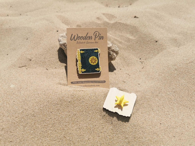 K&ouml;nnte beinhalten: Ein Holz-Pin in Form eines Buches mit goldenem Sonnendesign, pr&auml;sentiert auf einer Kraftpapierkarte. Der Pin ist dunkelgr&uuml;n mit goldenen Akzenten. Ein kleiner gelber sternf&ouml;rmiger Pin ist ebenfalls vorhanden.