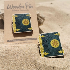 K&ouml;nnte beinhalten: Holz-Anstecknadel mit einer detaillierten Illustration eines Buches mit tealfarbenem Einband, goldenen Akzenten und einem zentralen Emblem. Die Anstecknadel liegt auf Sand mit einem Kartonr&uuml;cken.