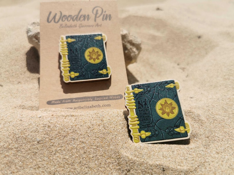 K&ouml;nnte beinhalten: Holz-Anstecknadel mit einer detaillierten Illustration eines Buches mit tealfarbenem Einband, goldenen Akzenten und einem zentralen Emblem. Die Anstecknadel liegt auf Sand mit einem Kartonr&uuml;cken.