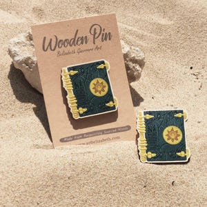 K&ouml;nnte beinhalten: Holz-Anstecknadel mit einem dunkelblauen Buchdesign mit goldenen Akzenten und einem zentralen Sonnenemblem. Die Anstecknadel wird auf einer braunen Karte mit dem Text "Wooden Pin" und "artelizabeth.com" pr&auml;sentiert. Eine zweite Anstecknadel ist ebenfalls sichtbar.