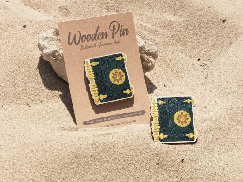 K&ouml;nnte beinhalten: Holz-Anstecknadel mit einem dunkelblauen Buchdesign mit goldenen Akzenten und einem zentralen Sonnenemblem. Die Anstecknadel wird auf einer braunen Karte mit dem Text "Wooden Pin" und "artelizabeth.com" pr&auml;sentiert. Eine zweite Anstecknadel ist ebenfalls sichtbar.