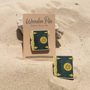 K&ouml;nnte beinhalten: Holz-Anstecknadel mit detailliertem Buchdesign und Sonnenemblem. Die Anstecknadel ist dunkelblaugr&uuml;n mit gelben Akzenten und wird auf einer braunen Karte mit dem Text "Wooden Pin" pr&auml;sentiert. Eine zweite Anstecknadel ist ebenfalls sichtbar.