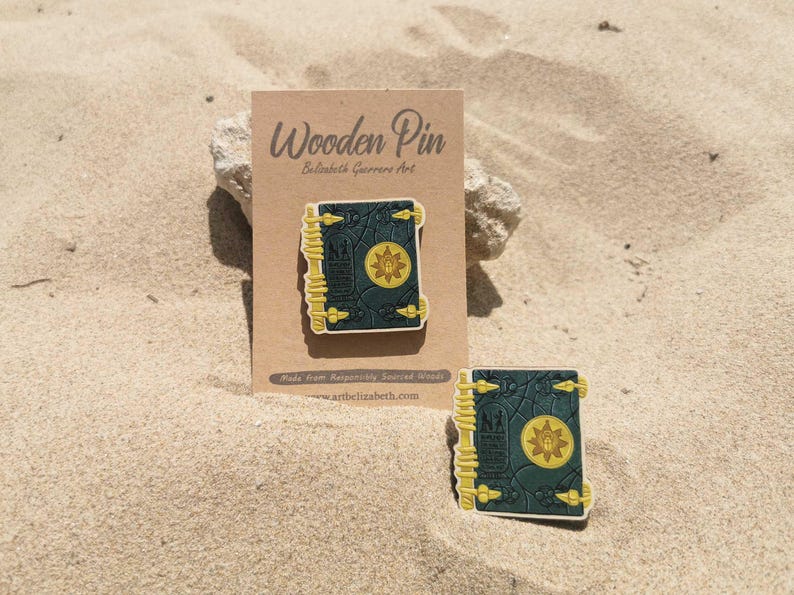 K&ouml;nnte beinhalten: Holz-Anstecknadel mit detailliertem Buchdesign und Sonnenemblem. Die Anstecknadel ist dunkelblaugr&uuml;n mit gelben Akzenten und wird auf einer braunen Karte mit dem Text "Wooden Pin" pr&auml;sentiert. Eine zweite Anstecknadel ist ebenfalls sichtbar.
