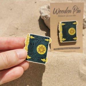 K&ouml;nnte beinhalten: Ein Holzpin in Buchform mit dunkelblauem Einband und goldenen Akzenten. Der Pin hat ein gelbes Emblem auf der Vorderseite und wird von einer Hand gehalten. Ein zweiter Pin wird auf einer Kartonr&uuml;ckseite gezeigt.