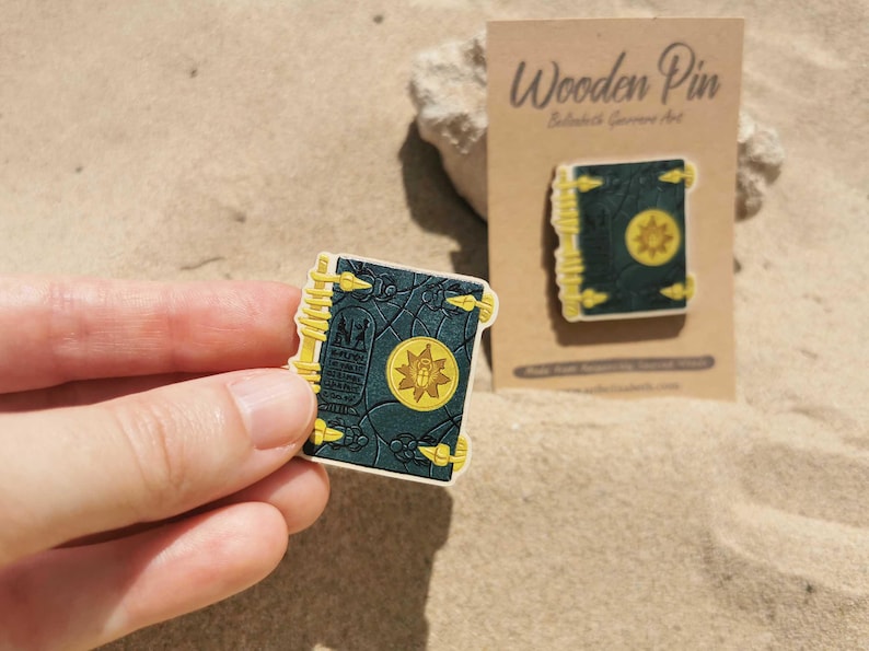 K&ouml;nnte beinhalten: Ein Holzpin in Buchform mit dunkelblauem Einband und goldenen Akzenten. Der Pin hat ein gelbes Emblem auf der Vorderseite und wird von einer Hand gehalten. Ein zweiter Pin wird auf einer Kartonr&uuml;ckseite gezeigt.