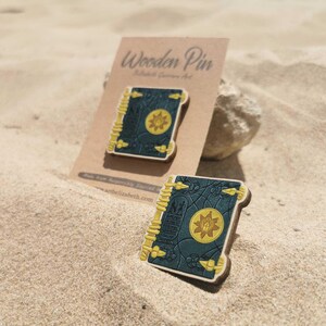 K&ouml;nnte beinhalten: Zwei Holzpins in Buchform mit dunkelblauem Einband und gelben Akzenten, auf Sand gelegt. Das Buchdesign zeigt ein goldenes Emblem. Die Pins werden auf einer braunen Karte mit der Aufschrift "Wooden Pin" pr&auml;sentiert.