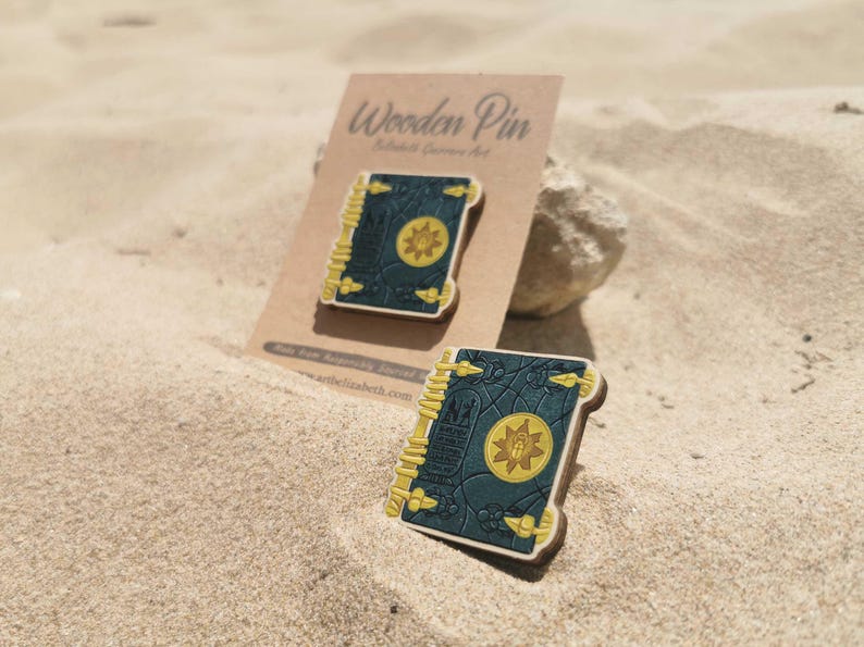 K&ouml;nnte beinhalten: Zwei Holzpins in Buchform mit dunkelblauem Einband und gelben Akzenten, auf Sand gelegt. Das Buchdesign zeigt ein goldenes Emblem. Die Pins werden auf einer braunen Karte mit der Aufschrift "Wooden Pin" pr&auml;sentiert.