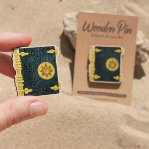 K&ouml;nnte beinhalten: Holz-Anstecknadel in Form eines Buches mit dunkelblauem Einband und goldenen Akzenten. Die Anstecknadel zeigt ein goldenes Emblem auf dem Einband und wird von einer Hand gehalten. Eine zweite Anstecknadel wird auf einer braunen Karte angezeigt.