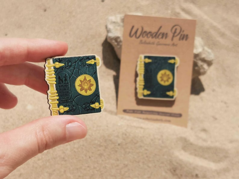 K&ouml;nnte beinhalten: Holz-Anstecknadel in Form eines Buches mit dunkelblauem Einband und goldenen Akzenten. Die Anstecknadel zeigt ein goldenes Emblem auf dem Einband und wird von einer Hand gehalten. Eine zweite Anstecknadel wird auf einer braunen Karte angezeigt.