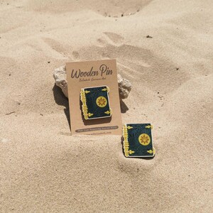 K&ouml;nnte beinhalten: Zwei Holzpins in Buchform mit dunkelblauem Einband und goldenen Akzenten. Ein Pin ist an einer braunen Karte mit dem Text "Wooden Pin" befestigt. Die Pins liegen auf einer sandigen Oberfl&auml;che.