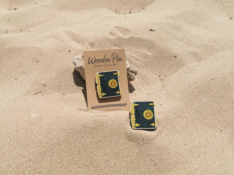 K&ouml;nnte beinhalten: Zwei Holzpins in Buchform mit dunkelblauem Einband und goldenen Akzenten. Ein Pin ist an einer braunen Karte mit dem Text "Wooden Pin" befestigt. Die Pins liegen auf einer sandigen Oberfl&auml;che.