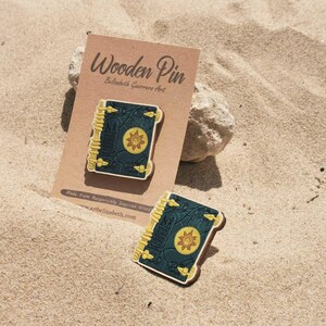 K&ouml;nnte beinhalten: Holz-Anstecknadel mit einem detaillierten Buchdesign mit dunkelgr&uuml;nem Einband, goldenen Akzenten und einem zentralen Emblem. Die Anstecknadel wird auf einer beigefarbenen Karte mit dem Text "Wooden Pin" und dem K&uuml;nstlernamen pr&auml;sentiert.