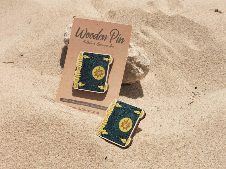 K&ouml;nnte beinhalten: Holz-Anstecknadel mit einem detaillierten Buchdesign mit dunkelgr&uuml;nem Einband, goldenen Akzenten und einem zentralen Emblem. Die Anstecknadel wird auf einer beigefarbenen Karte mit dem Text "Wooden Pin" und dem K&uuml;nstlernamen pr&auml;sentiert.