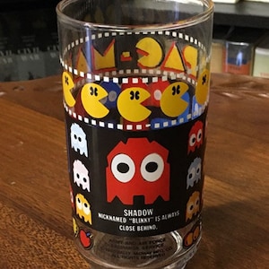 May include: A clear glass drinking glass with a black and yellow Pac-Man design. The glass features a red Pac-Man with the text "Shadow Nicknamed "Blinky" is always close behind."