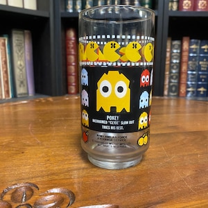 May include: A clear glass with a yellow Pac-Man character on a black background. The glass has a black rim and a white base. The text on the glass reads "POKEY NICKNAMED "CLYDE" SLOW BUT TRIES HIS BEST. ARMY AND AIR FORCE EXCHANGE SERVICE ©1980 BALLY MIDWAY MFG. CO. ALL RIGHTS RESERVED."