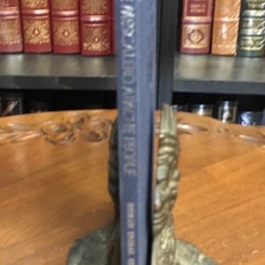 May include: A blue hardcover book titled "The Misadventures of People" by Peter Mayle. The book is standing upright on a bookend shaped like a person's head.
