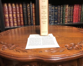 Könnte beinhalten: Ein Vintage-Hardcover-Buch mit dem Titel "Rip Van Winkle, The Legend of Sleepy Hollow and Other Stories" von Washington Irving. Das Buch ist Teil der Heritage-Serie und wird auf einem Holztisch mit aufwendigen Schnitzereien präsentiert.