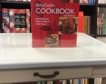 May include: A red Betty Crocker Cookbook with the text "Gift Edition" and "Everything You Need to Know to Cook Today". The cookbook is on a white table with a brown drawer handle.