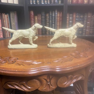 May include: Two white ceramic dog bookends in the shape of a hunting dog. The bookends are on a wooden table with intricate carvings.