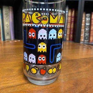 May include: A clear glass drinking glass with a black background and a Pac-Man design. The design features the Pac-Man character, ghosts, and a maze. The glass is decorated with yellow, red, blue, and white colors.