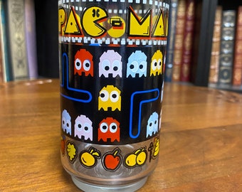 May include: A clear glass drinking glass with a black background and a Pac-Man design. The design features the Pac-Man character, ghosts, and a maze. The glass is decorated with yellow, red, blue, and white colors.