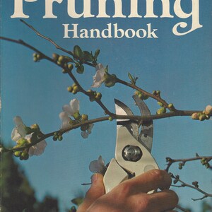 May include: The cover of the "Sunset Pruning Handbook" features the title in large white letters against a blue background. A hand uses pruning shears to cut a flowering branch. The book is a new edition.