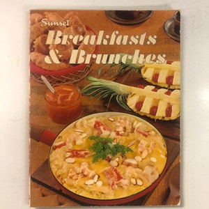 May include: A vintage cookbook titled "Sunset Breakfasts & Brunches" with a colorful cover featuring a skillet of shrimp and crab casserole, a bowl of fruit salad, and a basket of croissants.