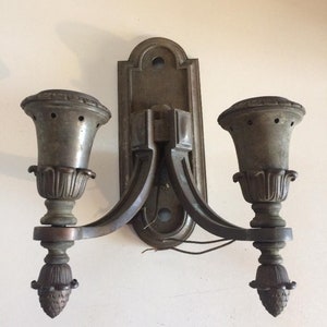 May include: A vintage brass wall sconce with two candle-style light fixtures. The sconce has a decorative design with a floral motif and a central plate.