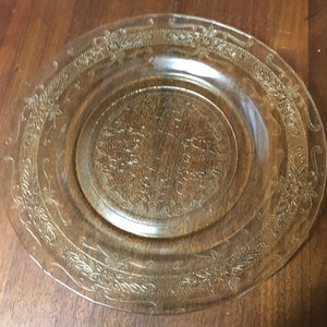 May include: A clear glass dinner plate with a delicate floral pattern etched into the surface. The plate has a slightly scalloped edge.