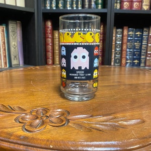 May include: A clear glass with a black and yellow Pac-Man design. The glass has a pink Pac-Man with the text "Speedy Nicknamed "Pinky" is pink and he's fast."  The text also includes "Army and Air Force Exchange Service Ted Bally Mowry Mfg. Co. All Rights Reserved."