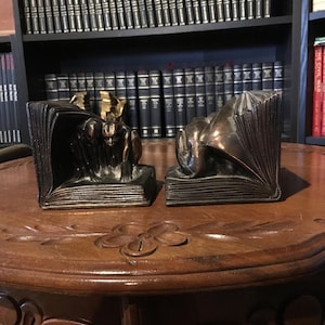 May include: A pair of bookend sculptures made of bronze. One bookend features a figure of a person reading a book, while the other bookend features a dragon reading a book. The bookends are sitting on a wooden table.