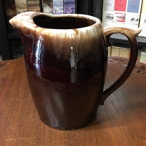 May include: A brown and white glazed ceramic pitcher with a wide mouth and a handle. The pitcher has a brown glaze with a white band around the top and a white glaze on the inside.