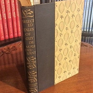 May include: A hardcover book titled "Selected Tales of Guy de Maupassant" with a dark spine and patterned cover. The book is standing upright on a wooden surface, with other books visible in the background.
