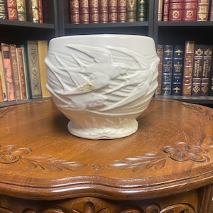 May include: A white ceramic planter with a raised design of birds and branches. The planter is sitting on a wooden table with intricate carvings.
