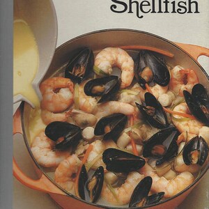 May include: A close-up of a pot filled with a seafood stew. The stew contains shrimp, mussels, and vegetables. The pot is orange and has a wooden handle. The text "THE GOOD COOK Shellfish" is printed on the top of the image.