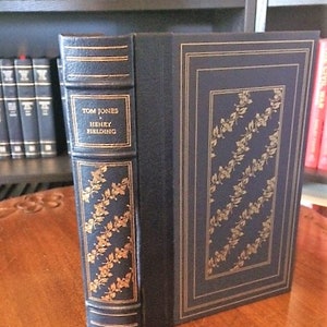 May include: A dark blue hardcover book with gold lettering on the spine. The title is "Tom Jones" by "Henry Fielding". The book has a gold patterned design on the front and back covers.