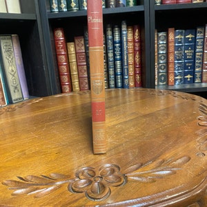 May include: A brown leather-bound book with gold lettering on the spine. The title is "Plotinus" and the book is part of the "Great Books" series. The book is sitting on a wooden table.