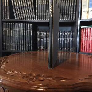 May include: A black hardcover book titled "COMUS" is standing upright on a wooden table. The book is surrounded by a bookshelf filled with other books.
