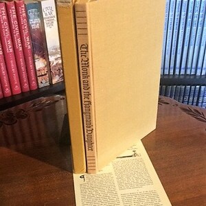 May include: A close-up of a book titled "The Monk and the Hangman's Daughter". The book is bound in brown leather and has gold lettering on the spine. The book is sitting on a table with other books in the background.