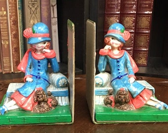 May include: A pair of bookends featuring a girl in a blue dress and hat, sitting on a white cushion with a brown poodle. The bookends are made of ceramic and have a green base.