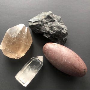 Peut inclure: Quatre cristaux et pierres naturelles sont disposés sur une surface sombre. Un grand cristal facetté brun clair est à côté d'un cristal clair et pointu. Une pierre gris foncé à texture rugueuse et une pierre lisse, ovale, brun rougeâtre complètent l'arrangement.