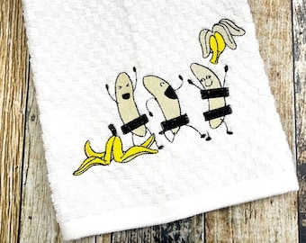 May include: White kitchen towel with an embroidered design of three bananas with faces and arms, wearing black censor bars over their middles. A banana peel is on the bottom left of the design.