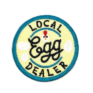 May include: Embroidered patch with a yellow center and a teal border. The patch reads "LOCAL EGG DEALER" in black script and features a small chicken illustration. Three white egg shapes are also included in the design.