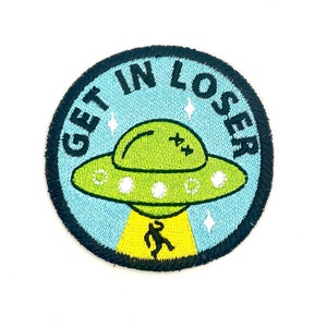 May include: A round, blue and green embroidered patch with a black outline. The patch features a cartoon UFO with a person running towards it. The text "GET IN LOSER" is written around the UFO.