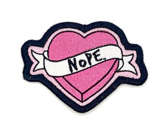 May include: A pink heart with a black outline and a white banner that says "NOPE" in black lettering. The heart and banner are embroidered on a black fabric patch.