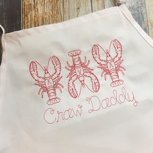 May include: White apron with red embroidered lobsters and the text "Crew Daddy".